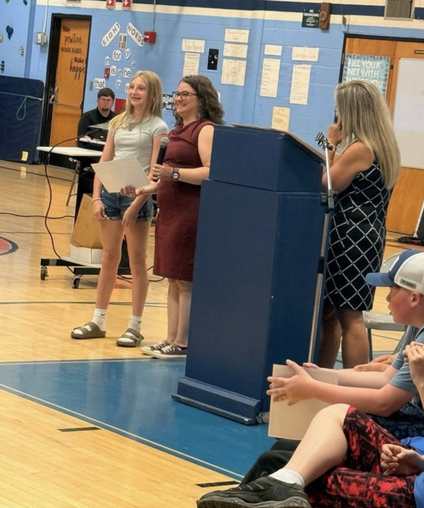A sixth-grade student stands proudly with a certificate while a teacher and staff member smile beside her during the celebration ceremony.