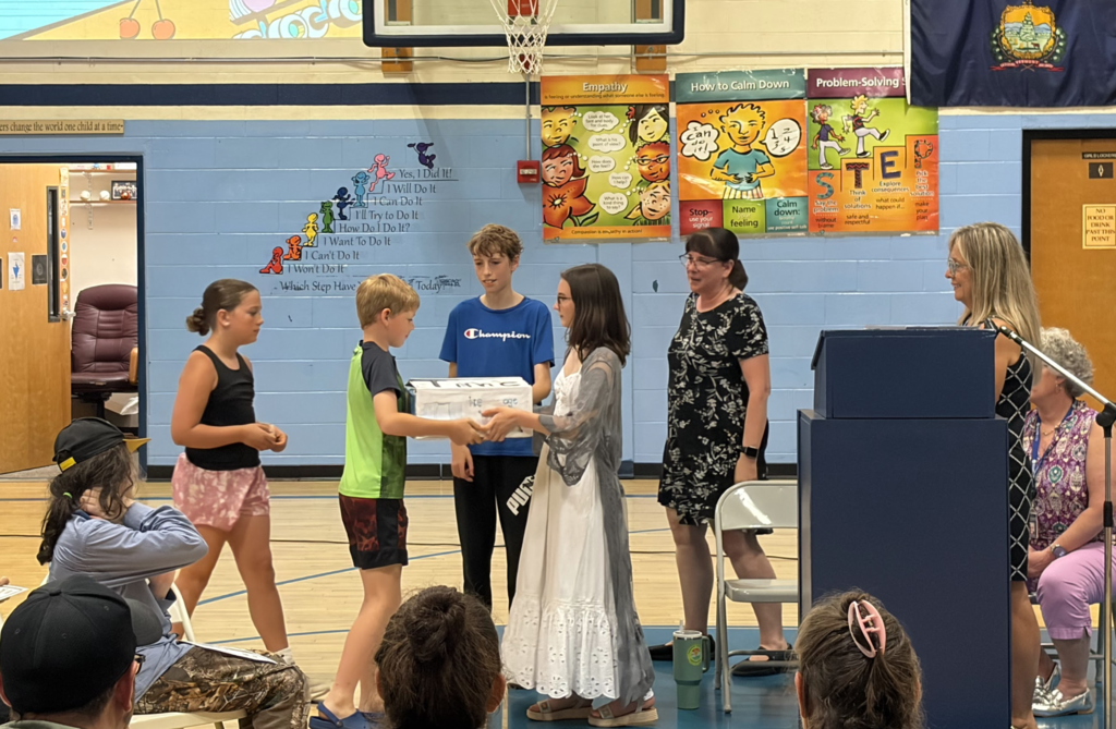 Several sixth-grade students participate in a recognition activity during the FHGS 6th Grade Celebration in the school gymnasium. Two students exchange a decorated memory box while staff members look on and families watch from the audience.