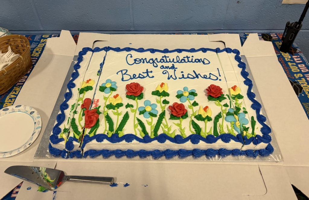 A decorated sheet cake with blue frosting and colorful flower designs sits on a table. The cake reads, “Congratulations and Best Wishes!” for the graduating sixth-grade class.