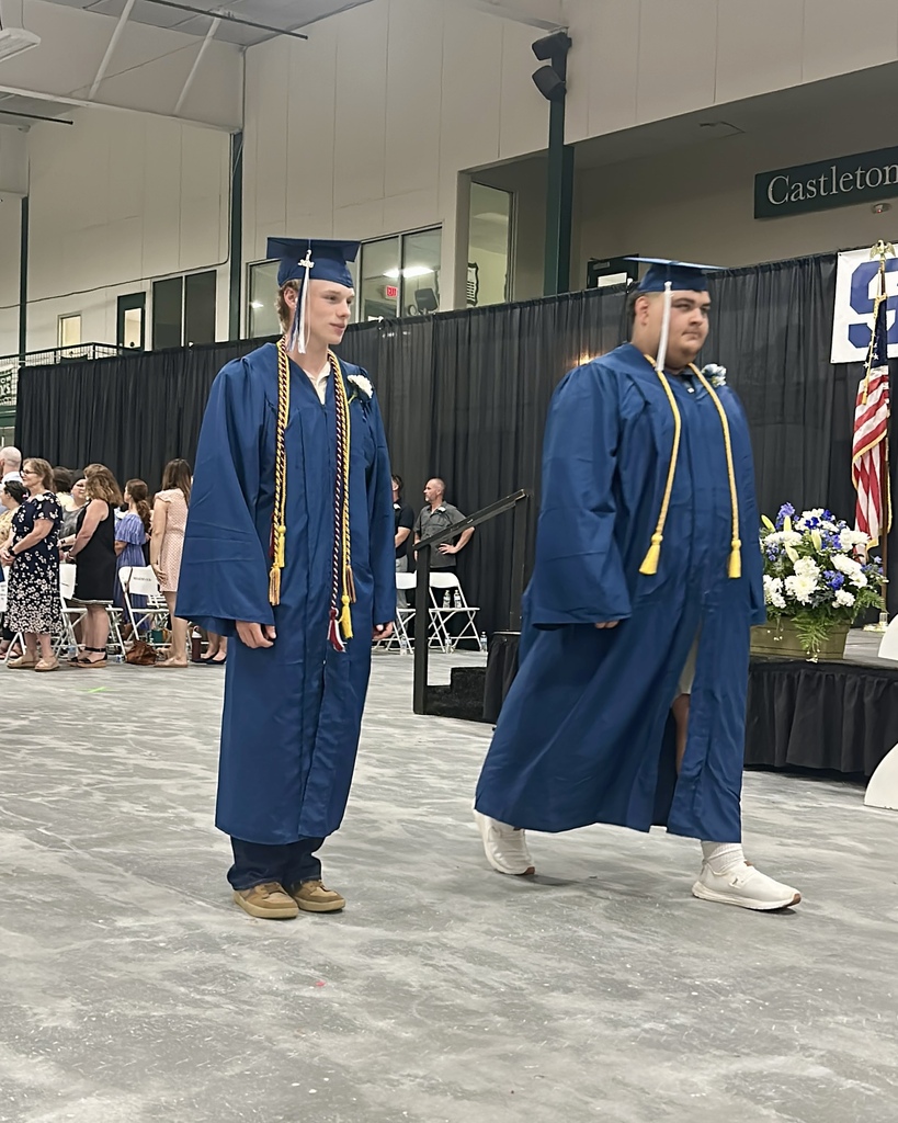 Two graduating seniors in blue caps and gowns walk across the stage during the commencement ceremony. Honor cords hang from their gowns as family and guests watch from the audience.