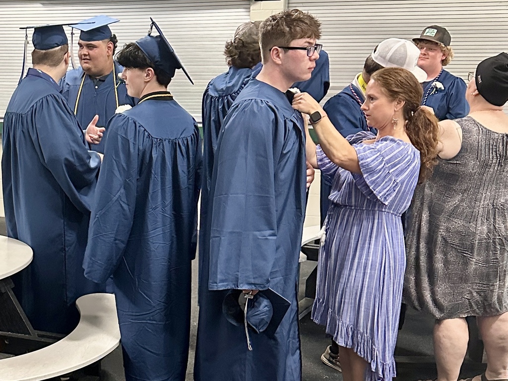 Several graduating seniors in blue caps and gowns gather backstage before commencement as family members help adjust boutonnieres and graduation attire.