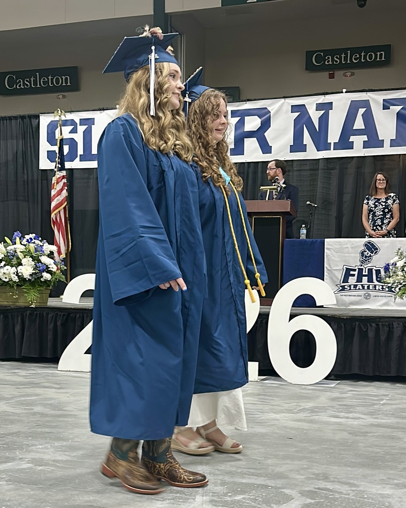 Two graduating seniors in blue caps and gowns walk across the stage during the commencement ceremony. Honor cords hang from their gowns as family and guests watch from the audience.