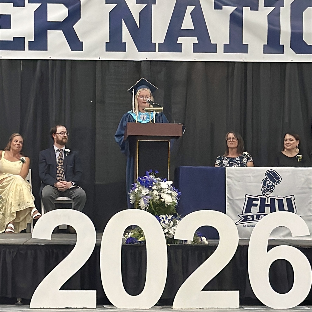 A graduating senior delivers a speech from the podium beneath the “Slater Nation” banner as faculty and administrators look on from the stage.