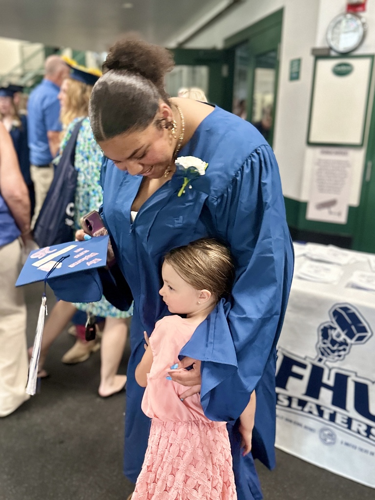 A graduating senior in a blue gown hugs a young child dressed in pink before the ceremony. The graduate smiles while holding a decorated graduation cap.