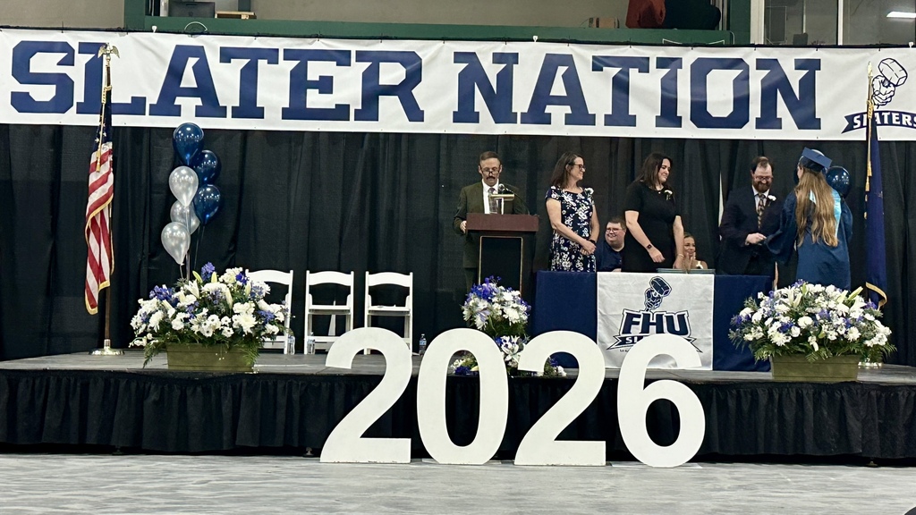 A graduate receives a diploma on stage during the Fair Haven Union High School commencement ceremony. The stage is decorated with blue-and-white floral arrangements, large “2026” numbers, and Slater Nation banners.