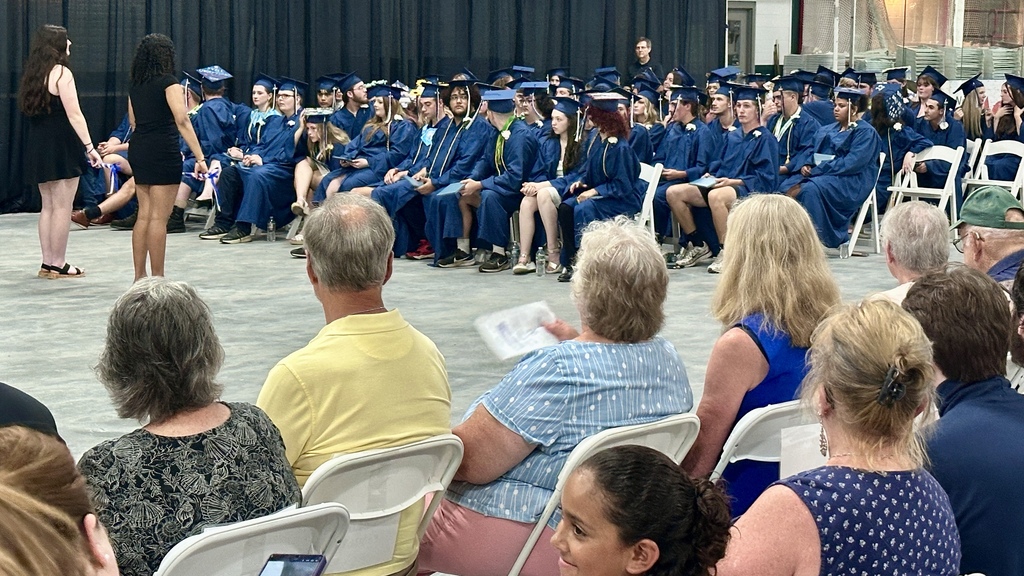 Graduates seated in rows of white chairs listen during the commencement ceremony while family members and guests watch from the audience.