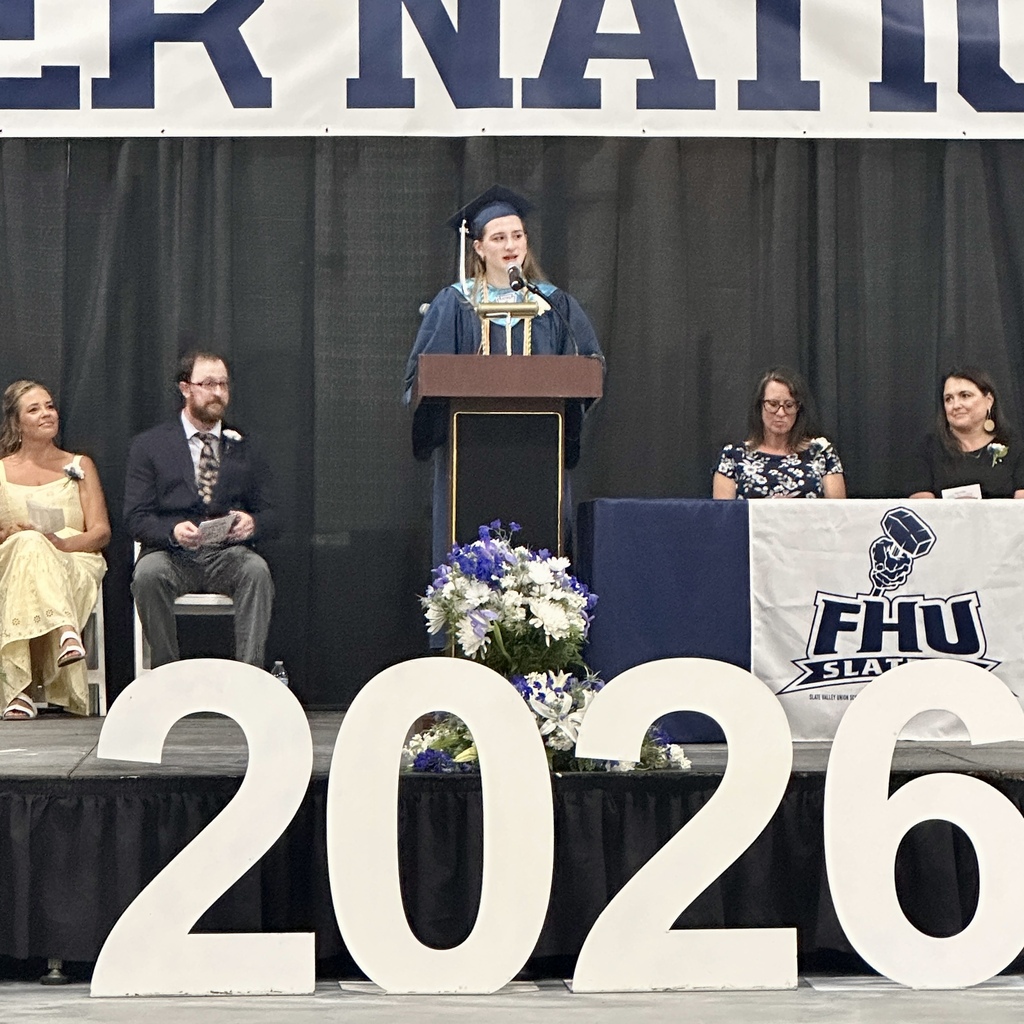 A graduating senior delivers a speech from the podium beneath the “Slater Nation” banner as faculty and administrators look on from the stage.