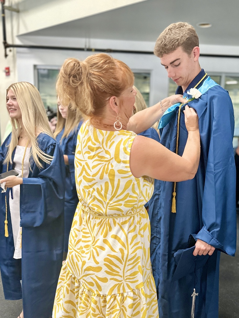 A family member pins a white flower boutonniere onto a graduating senior wearing a blue cap and gown before the Fair Haven Union High School graduation ceremony. Other graduates in blue gowns prepare nearby.
