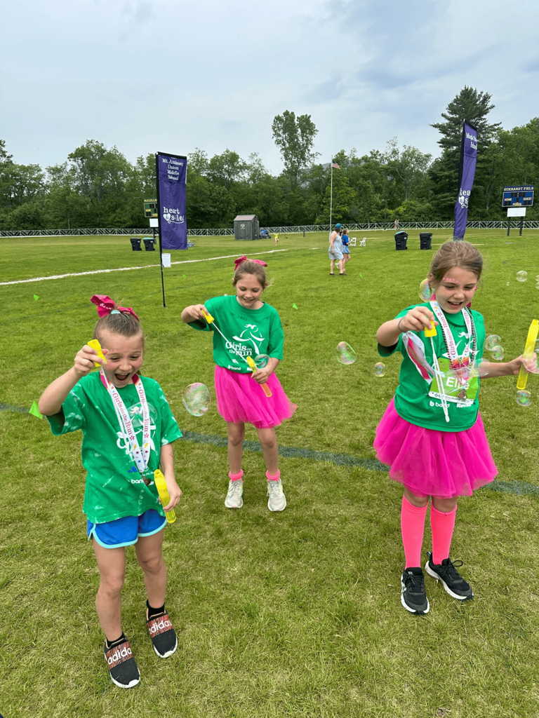 Three Girls on the Run participants wearing medals, green shirts, and pink tutus blow bubbles while standing on a grassy field after the 5K event.