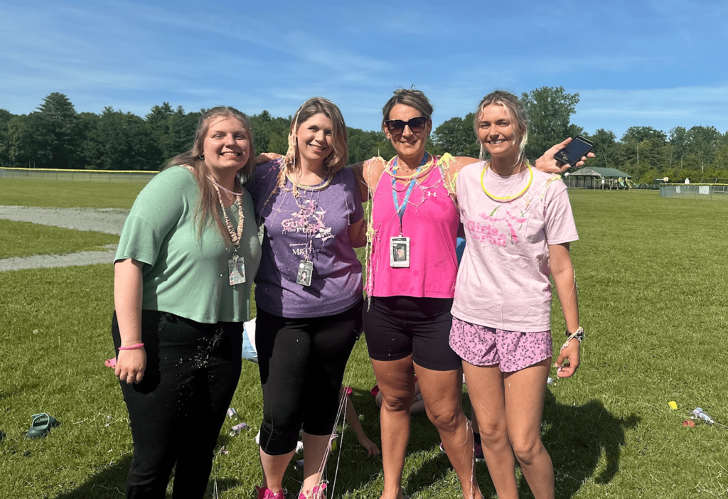 Four Girls on the Run coaches stand together on a sunny field, smiling after the season-ending celebration. Colorful silly string is draped across their shirts and shoulders.