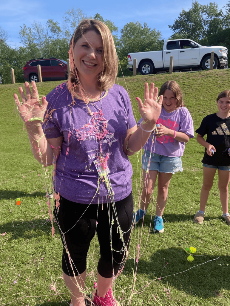 A Girls on the Run coach smiles while covered in colorful strands of silly string as students stand nearby laughing after the team's playful coach appreciation activity.