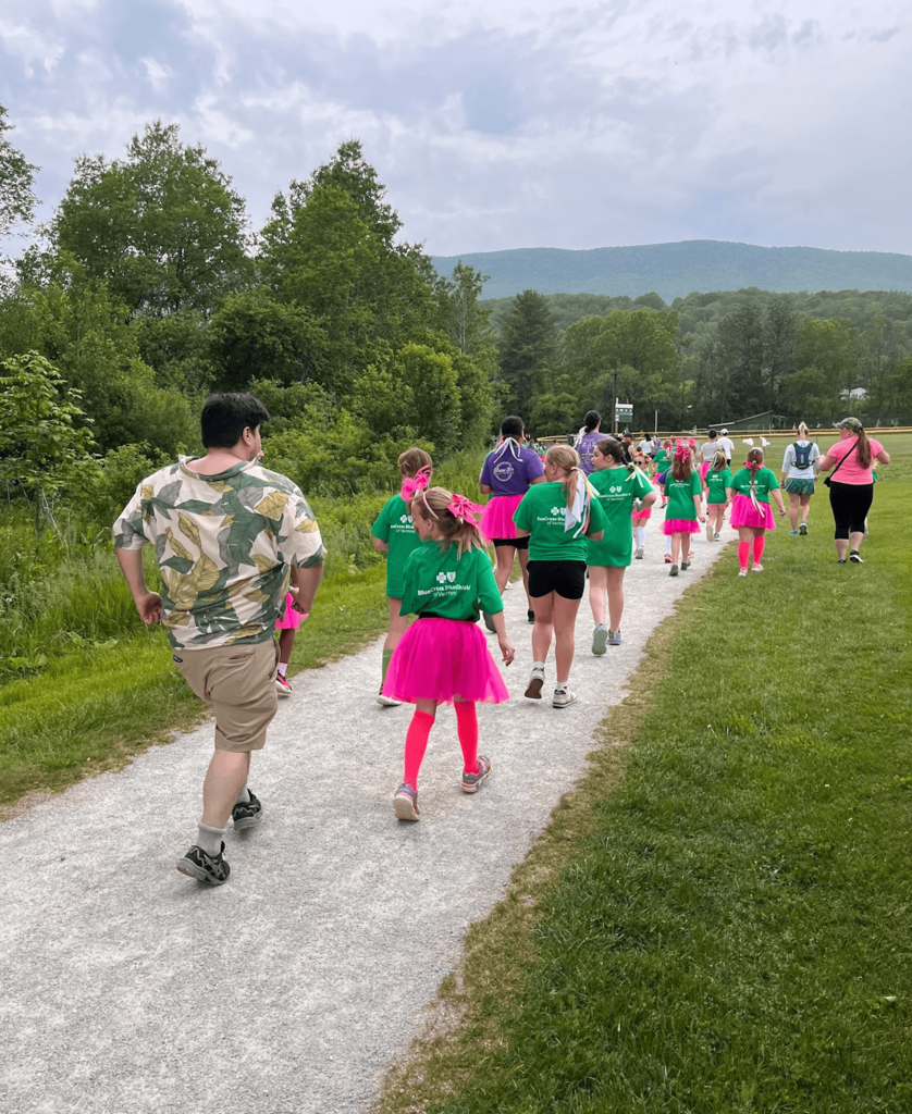 Participants and coaches walk together along a gravel path during the Girls on the Run 5K event, with mountains and trees visible in the background.