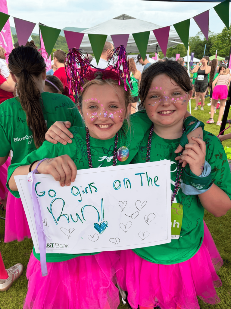 Two smiling Girls on the Run participants wearing green shirts, pink tutus, face paint, and festive headbands pose together while holding a handmade sign that reads, “Go girls on the Run!”