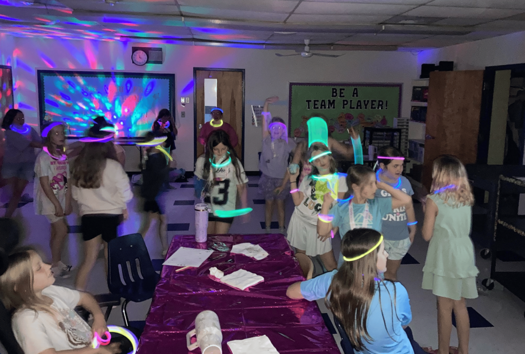 Students dance in a darkened classroom decorated with colorful glow sticks and party lights during a glow dance party celebration.