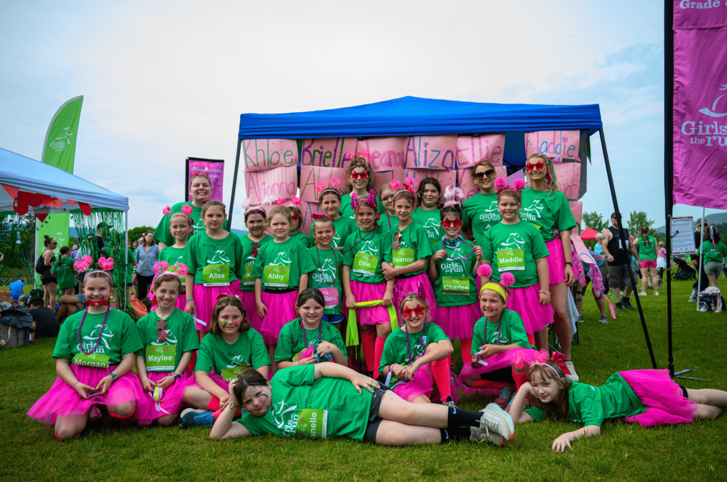 Large group of Girls on the Run participants and coaches pose together in front of a decorated team tent at the celebratory 5K. Students wear green Girls on the Run shirts, bright pink tutus, and colorful accessories.