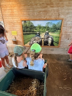 Two students sit in a wagon filled with hay while viewing a large farm display featuring a team of draft horses pulling farm equipment.