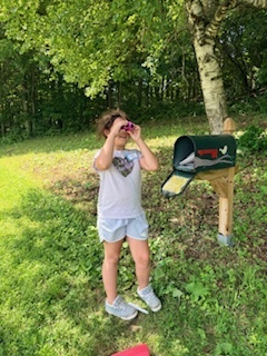A student stands outdoors near a decorative mailbox and uses binoculars to look across the farm landscape during an exploration activity.