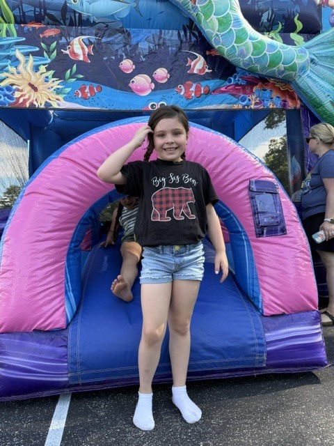 A young girl with braided hair smiles while standing at the bottom of a slide on an ocean-themed inflatable bounce house. She is wearing white socks, denim shorts, and a black shirt that reads "Big Sis Bear" with a red plaid bear graphic. Behind her, another child sits at the top of the slide, and the bounce house features colorful decorations of fish and a large mermaid tail.