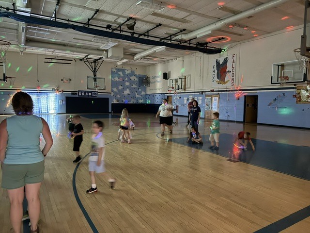 Several young children and adults gather on a wooden basketball court inside a school gymnasium for a glow party. The room is dimly lit with colorful green and red dance lights reflecting on the ceiling and walls. Many of the children are running and playing while wearing glowing necklaces or holding glow sticks. A banner on the wall features a falcon logo and reads "FHGS Falcons."