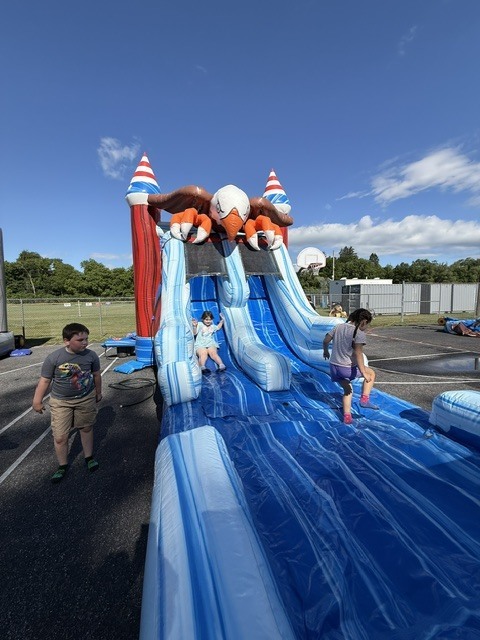 Children playing on a large, eagle-themed inflatable slide set up outdoors on an asphalt lot under a clear blue sky. One young girl smiles as she slides down the blue and white striped chute, another girl stands near the landing , and a young boy watches from the side.