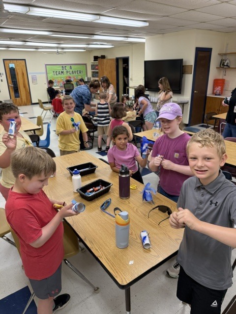 Elementary students gathered around classroom tables participating in a hands-on activity, smiling and showing completed craft projects while working together in small groups.