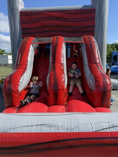Two young boys sit side-by-side at the bottom of parallel slides on a large red and grey inflatable obstacle course structure outdoors. The boy on the left is wearing a grey t-shirt and black shorts with his hands slightly raised, while the boy on the right wears a graphic t-shirt and khaki shorts. A bright blue sky with light clouds is visible above the top of the inflatable.