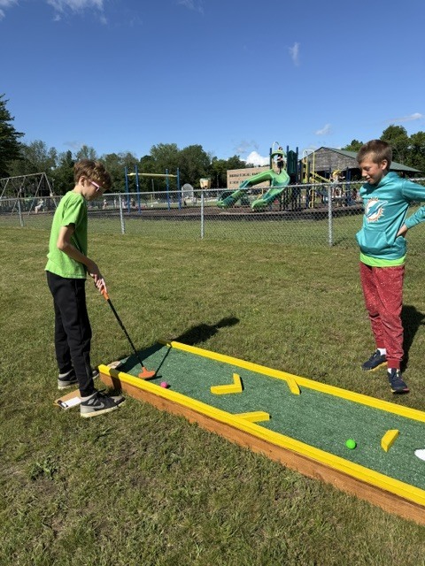 Two young boys play a game of mini-golf outdoors on a grassy lawn under a clear blue sky. One boy in a bright green t-shirt and black pants holds a putter, preparing to hit a pink golf ball on a narrow, portable mini-golf lane with yellow borders. Another boy wearing a teal Miami Dolphins sweatshirt and red pants stands by the lane, watching the play. A fenced-in school playground with slides and swings is visible in the background.
