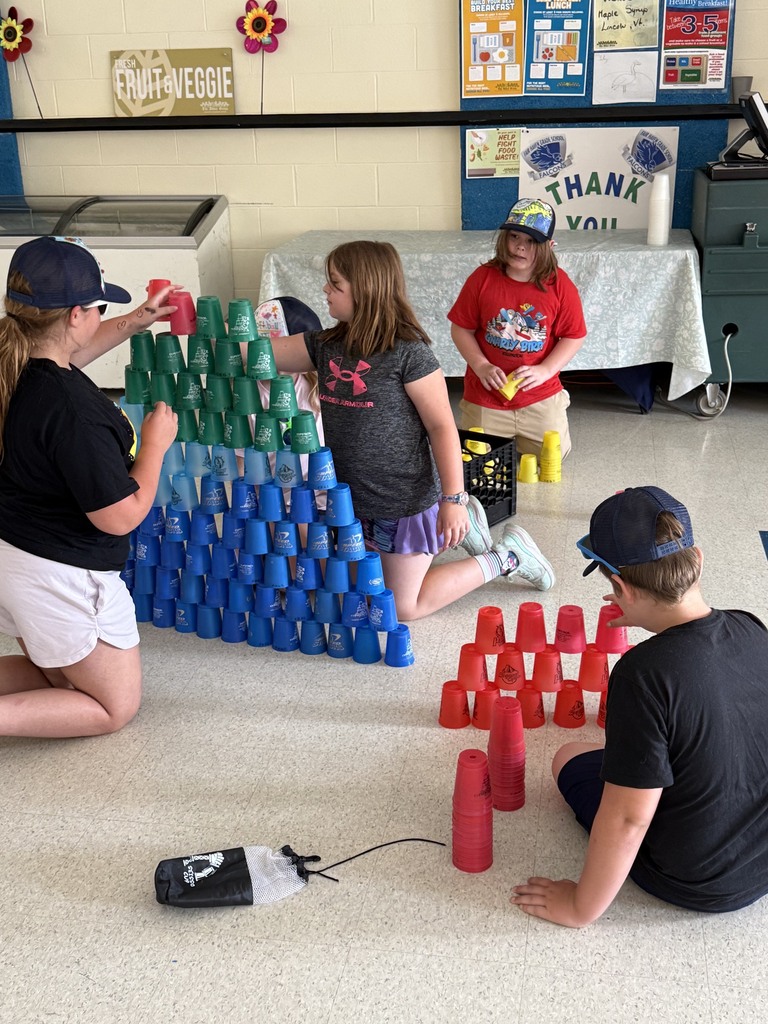 Four children sit on a school cafeteria floor building large pyramids out of colorful plastic stacking cups. One group of children collaborates on a large green and blue cup tower, while another boy constructs a smaller red cup pyramid nearby.