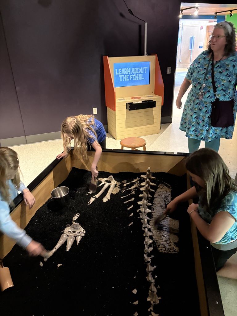 A high-angle shot of an indoor museum fossil dig exhibit. Three children are gathered around a large, rectangular wooden bin filled with black sand, using brushes to uncover a white replica skeleton of a dinosaur's spine and limbs. An adult woman in a blue patterned dress and teal leggings stands nearby watching, and a kiosk in the background displays a blue screen reading "Learn About the Fossil."