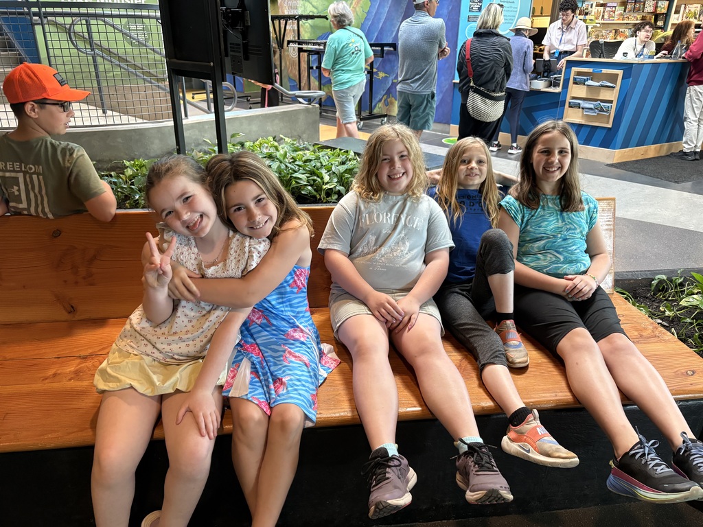 A wide shot of five young girls sitting side-by-side on a long, polished wooden bench inside a museum lobby. The two girls on the far left are hugging tightly and smiling, with one making a peace sign. The other three girls sit in a row next to them, smiling happily. In the background, other museum visitors, a service desk, and an exhibit staircase are visible.