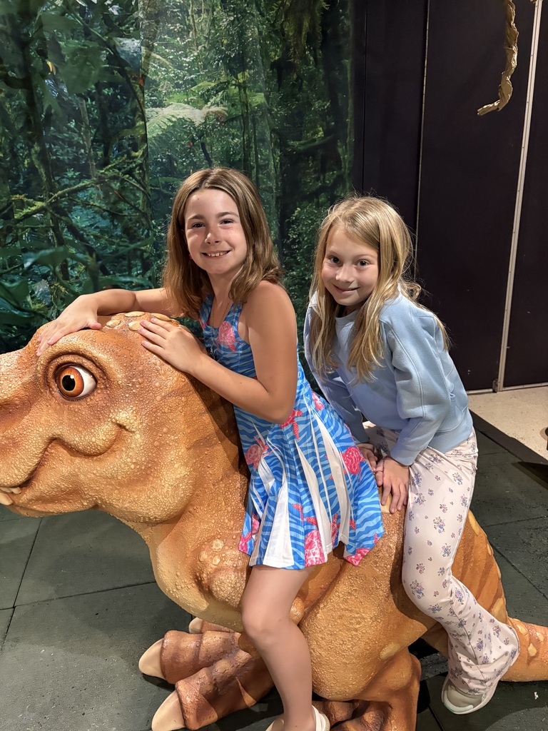 A medium shot of two smiling young girls riding a large, brown toy dinosaur prop inside a museum or exhibit. The girl in front is wearing a sleeveless blue dress with a pink turtle pattern, holding onto the dinosaur's head. The girl behind her is wearing a light blue sweatshirt and white floral-patterned pants, smiling warmly at the camera. The background features a green, dense jungle backdrop.
