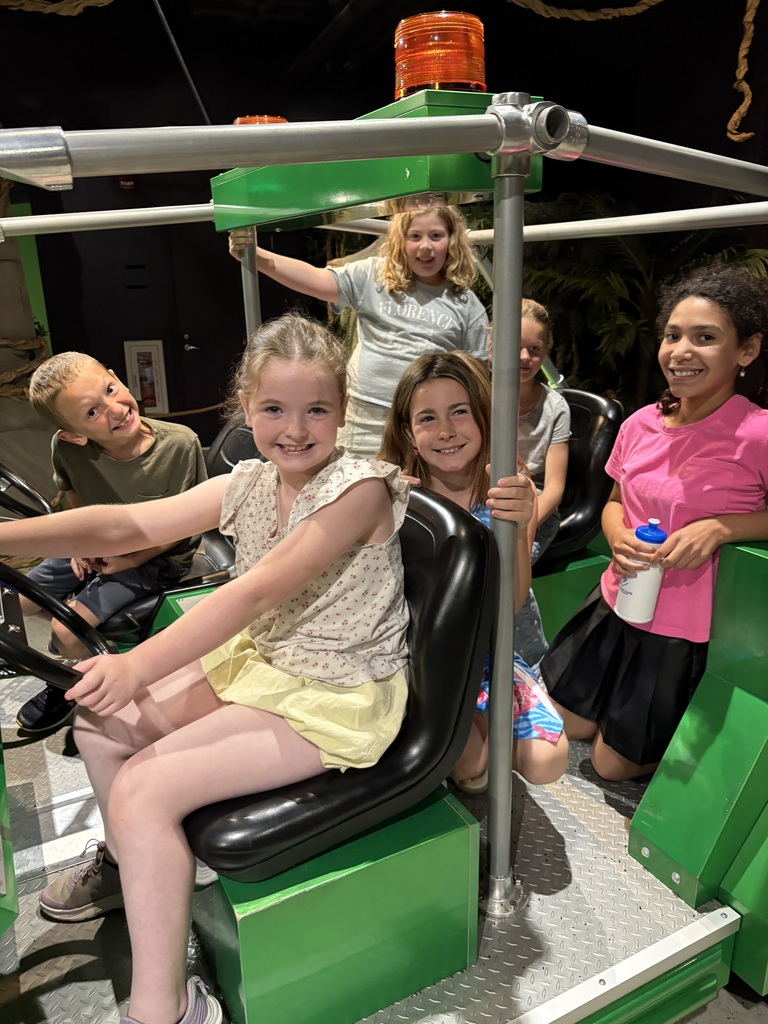 A medium shot similar to the previous image, showing two young girls posing on a brown toy dinosaur prop. The girl in the front wears a blue dress with a pink turtle pattern and smiles directly at the camera, while the girl behind her, wearing a light blue sweatshirt, looks off-to the side with a gentle smile. They are set against a lush, green jungle wall backdrop.