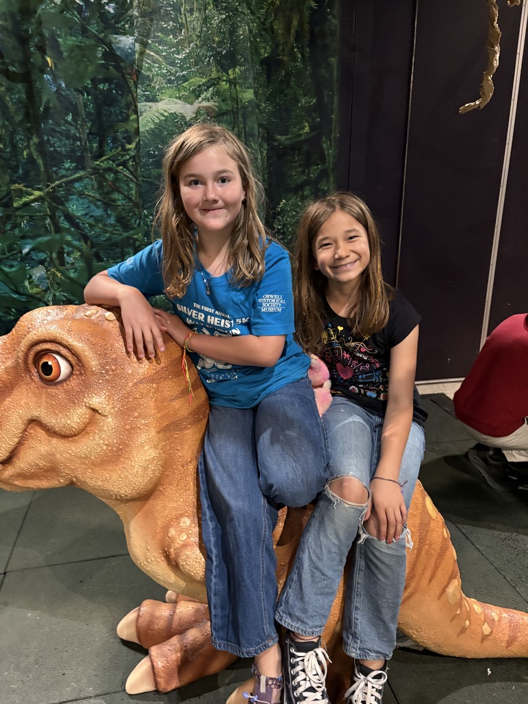 A medium shot of two smiling young girls riding a large, brown toy dinosaur prop inside a museum or exhibit. The girl in front is wearing a sleeveless blue dress with a pink turtle pattern, holding onto the dinosaur's head. The girl behind her is wearing a light blue sweatshirt and white floral-patterned pants, smiling warmly at the camera. The background features a green, dense jungle backdrop.