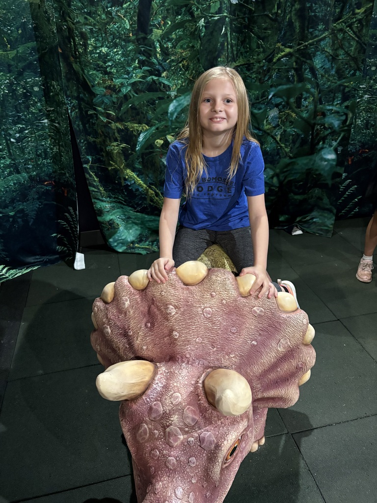 A smiling girl in a blue marbled t-shirt sits on the back of a large, realistic model dinosaur with textured orange skin and large eyes. In the background, a fossil dig pit with a dinosaur skull and other prehistoric models are visible.