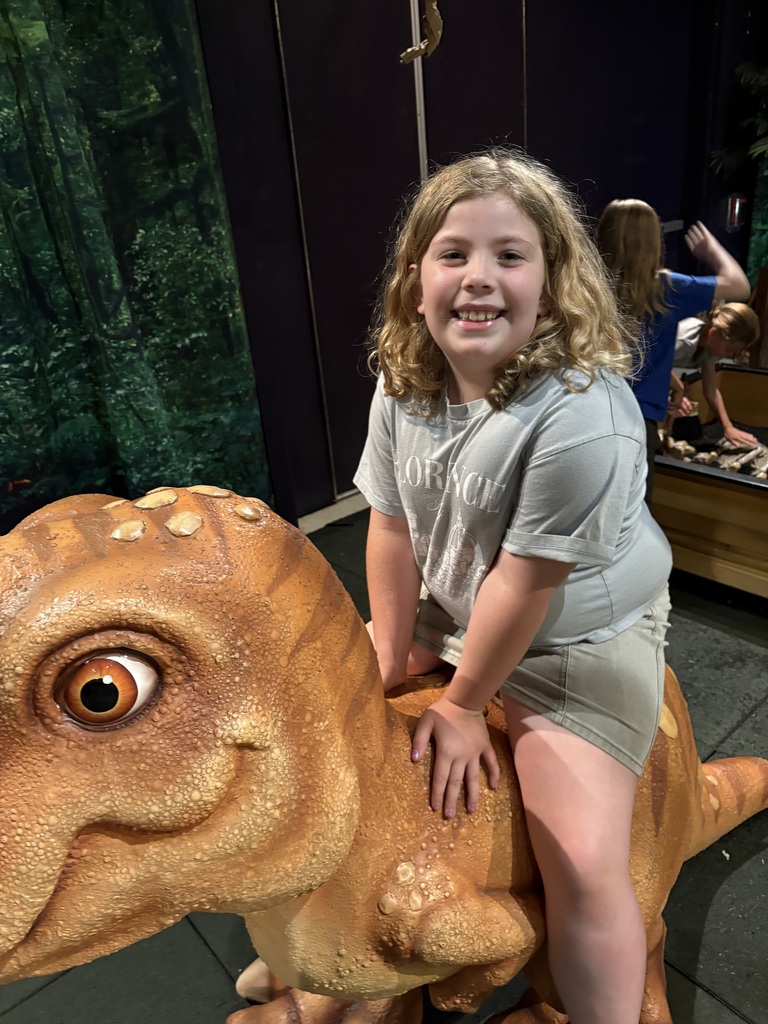A smiling girl in a blue marbled t-shirt sits on the back of a large, realistic model dinosaur with textured orange skin and large eyes. In the background, a fossil dig pit with a dinosaur skull and other prehistoric models are visible.
