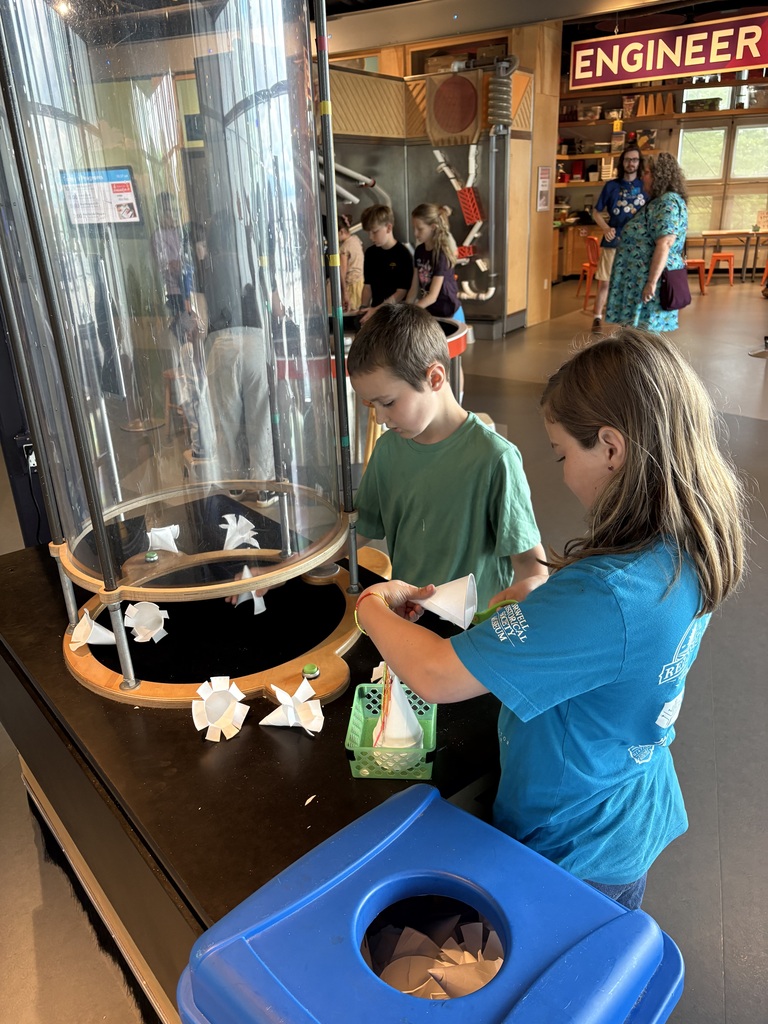 A young boy in a green shirt and a girl in a bright blue t-shirt construct paper cones at an interactive science museum exhibit. They stand in front of a tall, clear vertical wind tunnel tube where other paper structures are being tested. A blue bin filled with discarded paper cones sits in the foreground.