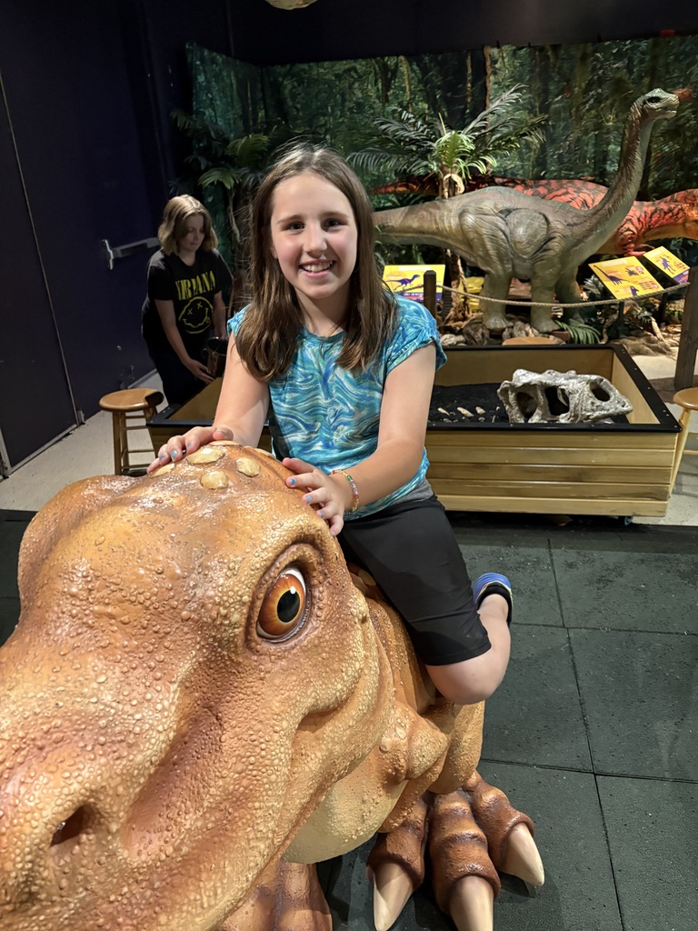 A young girl with long blonde hair and a blue t-shirt smiles while sitting astride a model Triceratops dinosaur. She rests her hands on the dinosaur's bony frill.