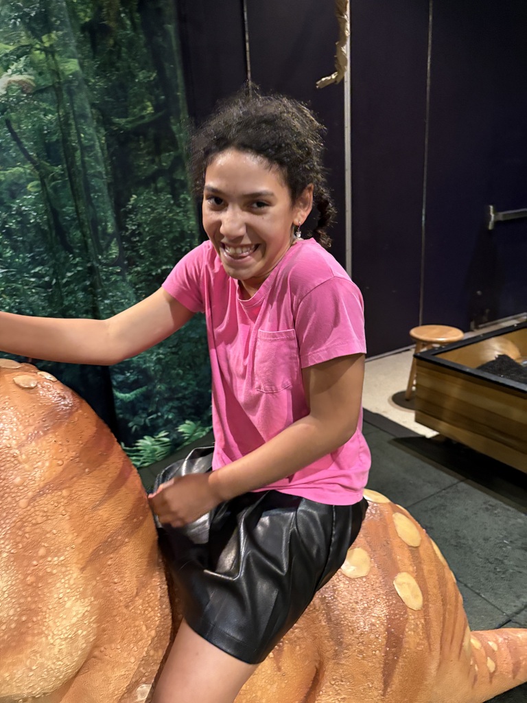 A teenager in a bright pink t-shirt and black shorts laughs while sitting on the back of a large orange dinosaur model against a dark wall and jungle backdrop.