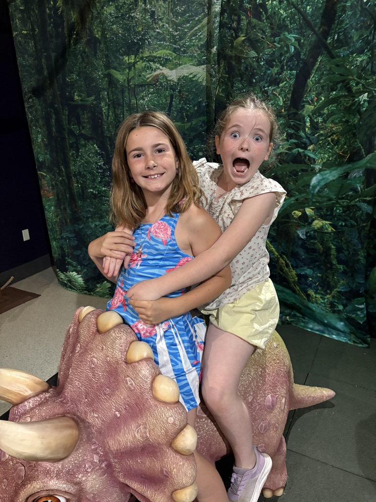 Two young girls smile playfully while riding together on a life-sized, pinkish-purple model of a Triceratops dinosaur. The girl in the back has a wide-eyed, comical expression with her mouth open in a mock scream, while the girl in front wears a blue patterned dress and smiles warmly. The background features a dense jungle backdrop.