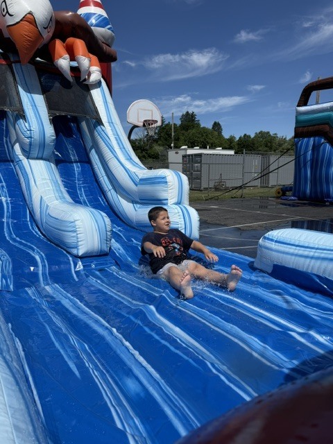 A young boy splashing down a large, blue and white inflatable water slide on a sunny day. He is sliding on his back with his feet forward. In the background of image_eba260.jpg, a basketball hoop, fence, and a clear blue sky with light clouds are visible.