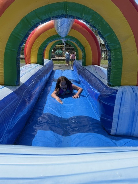 A young girl smiling and sliding on her stomach down a wet, blue inflatable slip-and-slide. The slide features a tunnel of large, colorful rainbow arches stretching over the track.