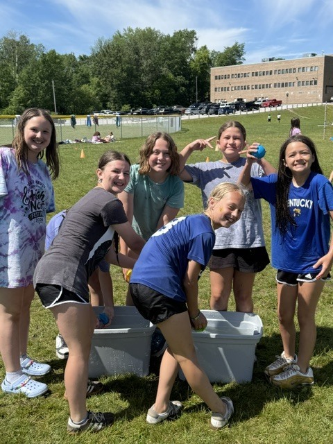 A group of six smiling young girls playing a water game outside on a sunny grassy field. Two large grey plastic bins filled with water sit on the grass in front of them as they laugh and pose for the camera. A large brick building and a parking lot are visible on a hill in the background