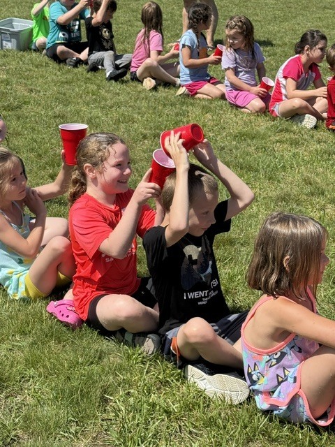 Here is a brief, ADA-compliant alt text description for the image: Alt Text: A line of young children sitting on a grassy field playing a water relay game with red plastic cups on a sunny day. In the foreground, a smiling girl in a red shirt pours water from her cup into a cup held over the head of the boy sitting in front of her. Another line of children playing the same game is visible in the background