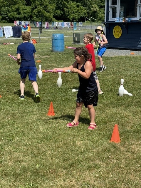 A group of young children playing an outdoor game with water blasters on a sunny, grassy field. In the foreground, a girl in a black tank top and shorts smiles as she holds a pink water toy. Orange cones and white plastic bowling pins are spread across the grass.