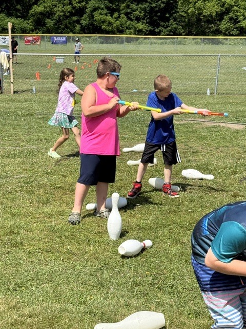 Two boys stand on a grassy field playing a game with long tube-style water blasters on a sunny day. White plastic bowling pins are scattered on the grass around their feet, with some standing upright and others knocked over. In the background of image_eba201.jpg, another child runs across the field near a chain-link fence.