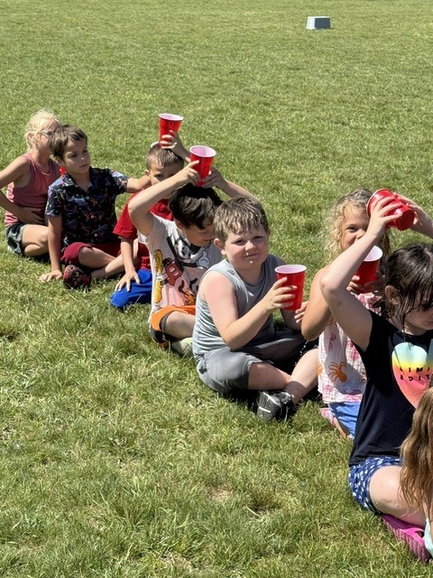 Here is a brief, ADA-compliant alt text description for the image: Alt Text: A line of young children sitting cross-legged on a sunny, grassy field, playing a water game with red plastic cups. Some children are holding the cups over their heads to pass water backward, while others hold them in front of them, smiling at the camera.