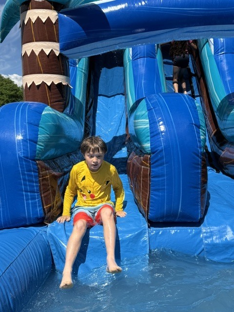 A young boy wearing a yellow Pikachu rash guard and red swim trunks sits at the bottom edge of a large, blue inflatable water slide, preparing to splash into the pool below. Another child is visible climbing up the stairs of the inflatable structure in the background
