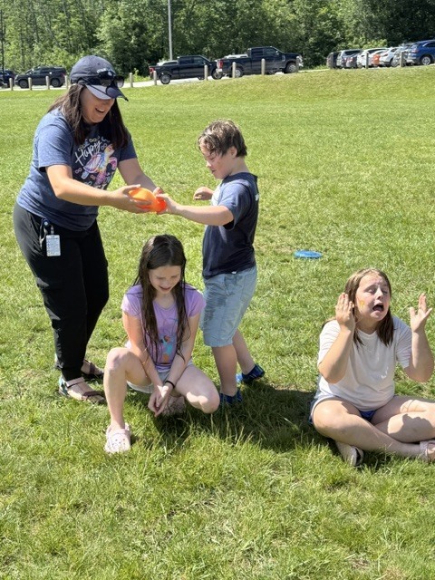 An adult wearing a baseball cap and a group of three children are playing a game with an orange water balloon on a sunny, grassy field. One boy stands up to catch or pop the balloon held by the adult, while a girl crouches down beside him and another girl sits cross-legged on the grass with her hands up in mid-reaction.