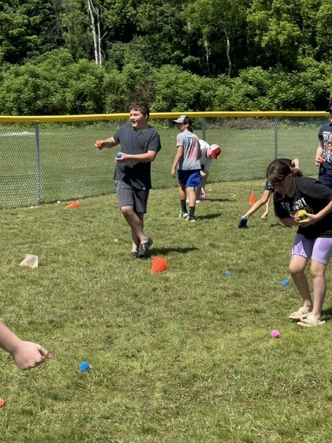 Here is a brief, ADA-compliant alt text description for the image: Alt Text: Several children play an outdoor game on a sunny, grassy field. In the center, a boy in a dark t-shirt and shorts looks off to the side while holding small, colorful balls. To the right, a girl leans forward to pick up or hold a ball, while other children bend down across the lawn in the background near orange cones. A chain-link fence topped with a yellow bar runs across the field, with a dense backdrop of green trees