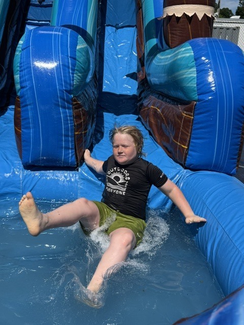 A boy wearing a black t-shirt and green shorts splashes into the shallow pool at the bottom of a large, blue inflatable water slide. His arms are spread wide and his feet are elevated as he slides into the water on a sunny day